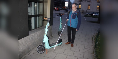 Am Laimer Platz erklärt Stefan Unterstraßer, warum auf dem Gehweg zurückgelassene Roller für ihn gefährliche Stolperfallen sind. Zudem machen es ihm die Scooter wie hier unmöglich, sich mit seinem Taststock an der Kante entlang des U-Bahn-Aufzugs und des Gehweg zu orientieren. (Foto: job)